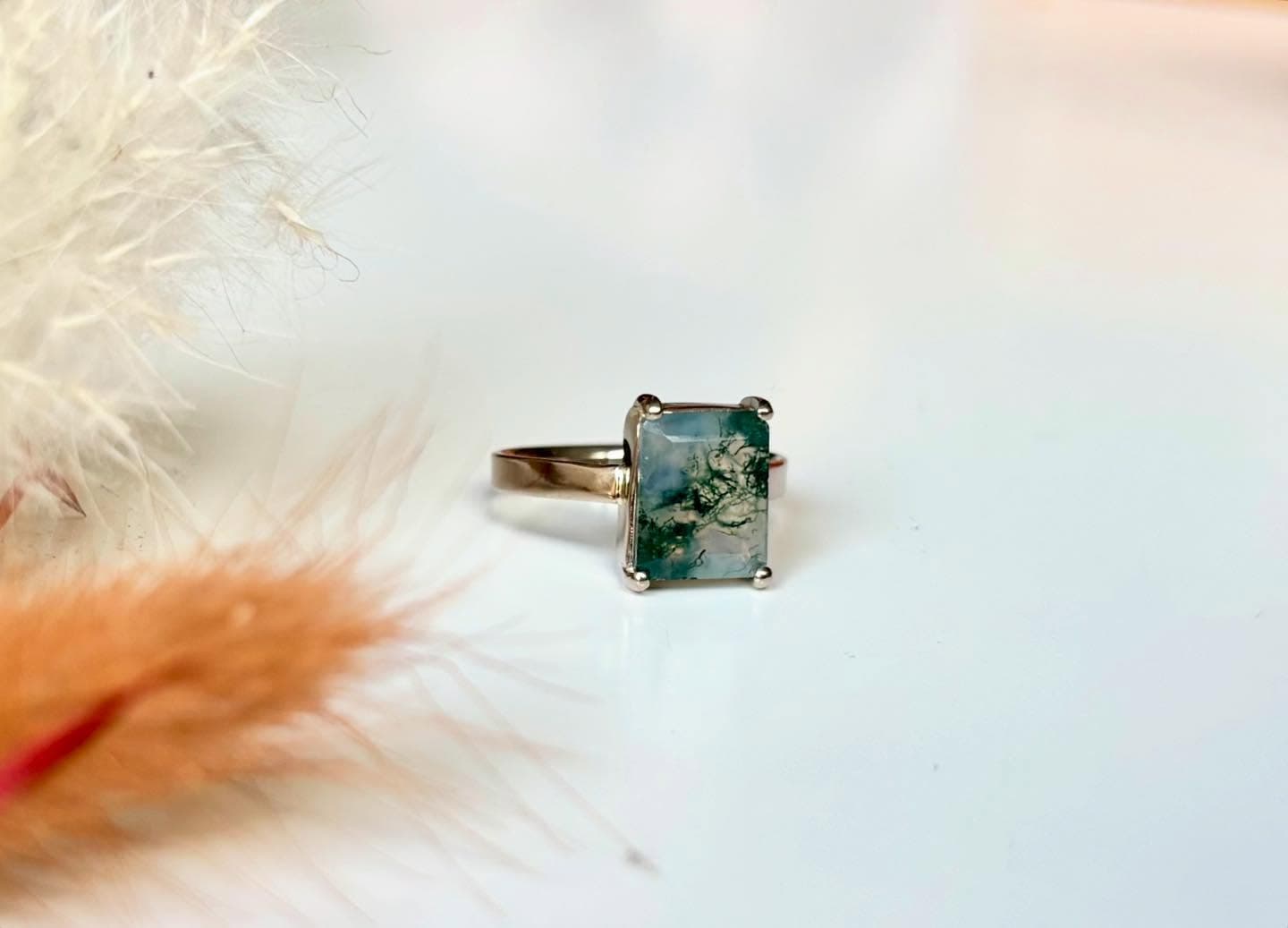 Gouden ring met vierkante groene steen (mogelijk mosagaat) op lichte ondergrond met veren